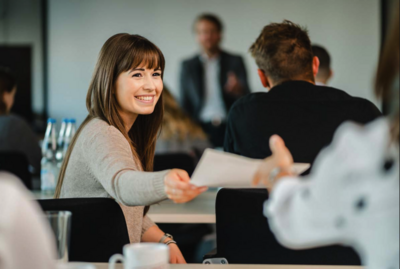 Frau reicht im Seminarraum ein Blatt nach hinten durch